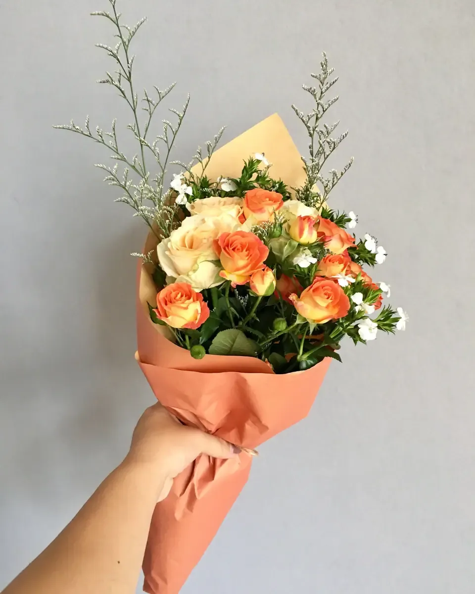 Bó hồng cam đào cùng cúc nhỏ gói giấy cam đất được cầm trên tay