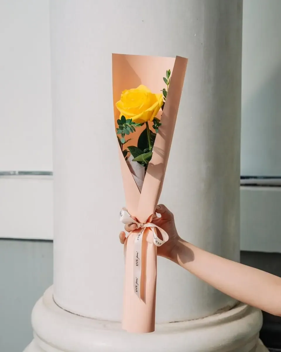 Một bông hồng vàng gói giấy hồng cam dạng nón, buộc ruy băng mảnh, được cầm trước cột trắng