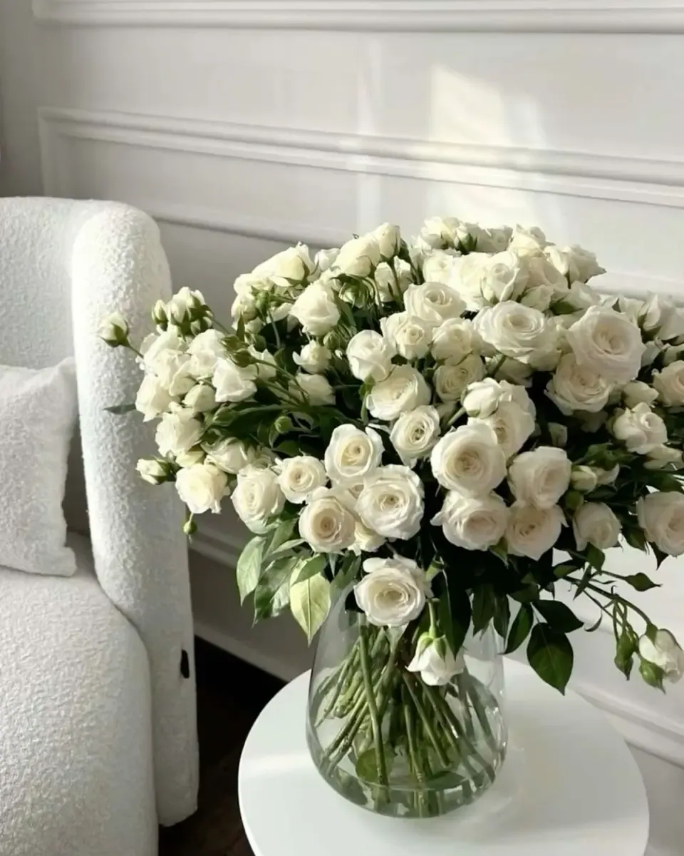 Bình thủy tinh cắm hoa hồng trắng nhỏ đặt cạnh ghế bành trắng trước tường ốp sáng