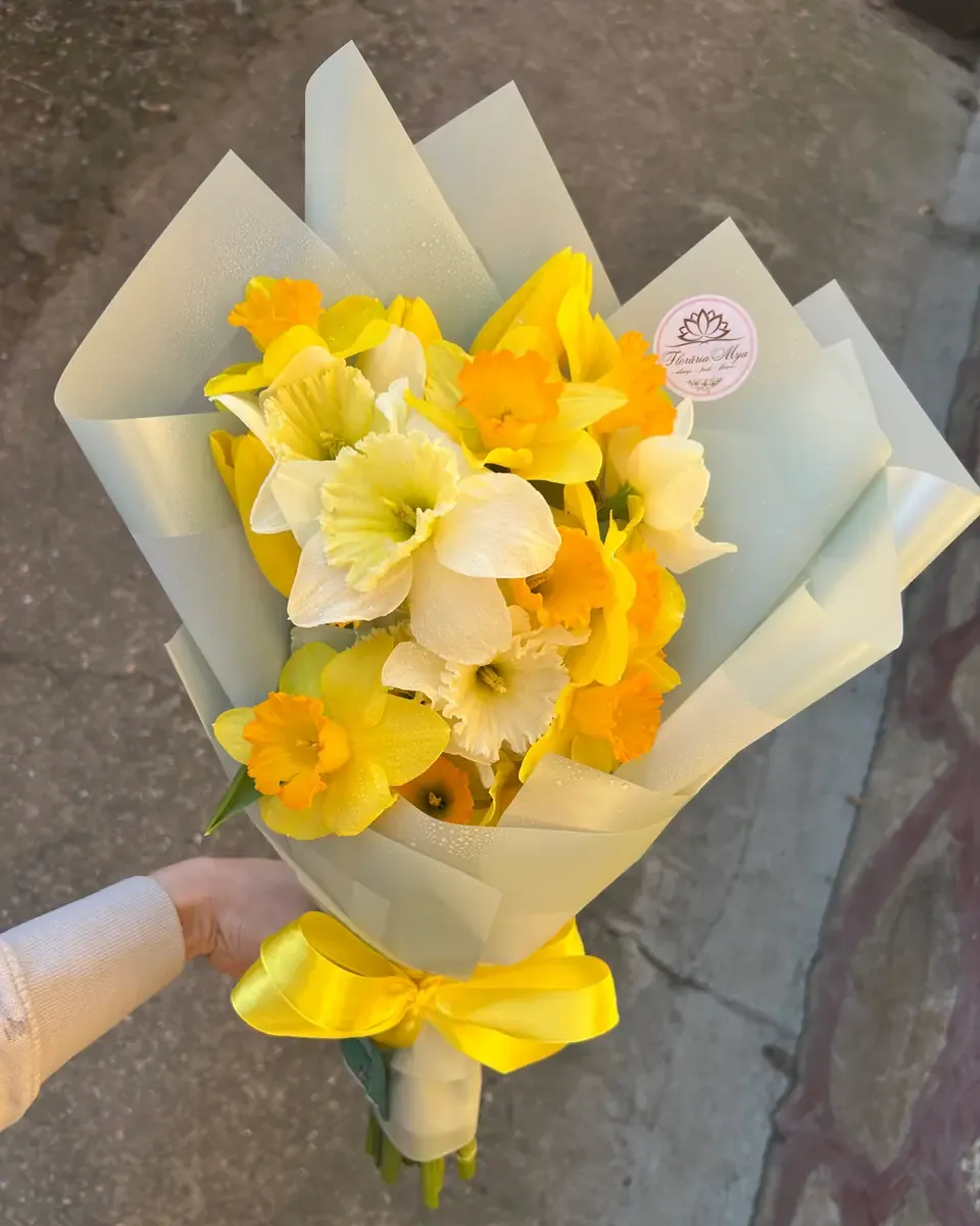 Bó thủy tiên vàng và cam gói giấy xanh nhạt, buộc ruy băng vàng, một tay cầm bó hoa