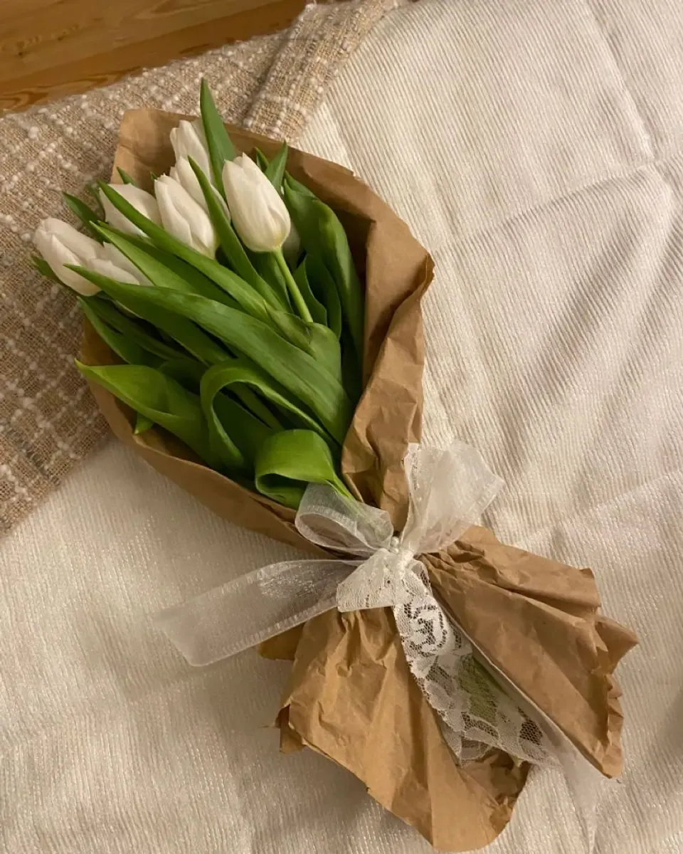 Bó tulip trắng gói giấy kraft nâu, buộc ruy băng ren kem, đặt trên tấm vải dệt sáng