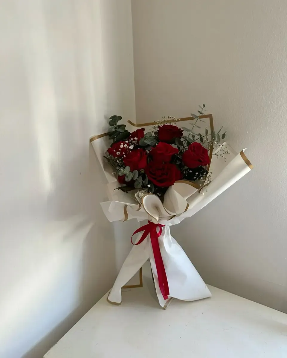 Bó hoa hồng đỏ nhỏ gói giấy kem và trắng, buộc ruy băng đỏ, đặt ở góc phòng sáng