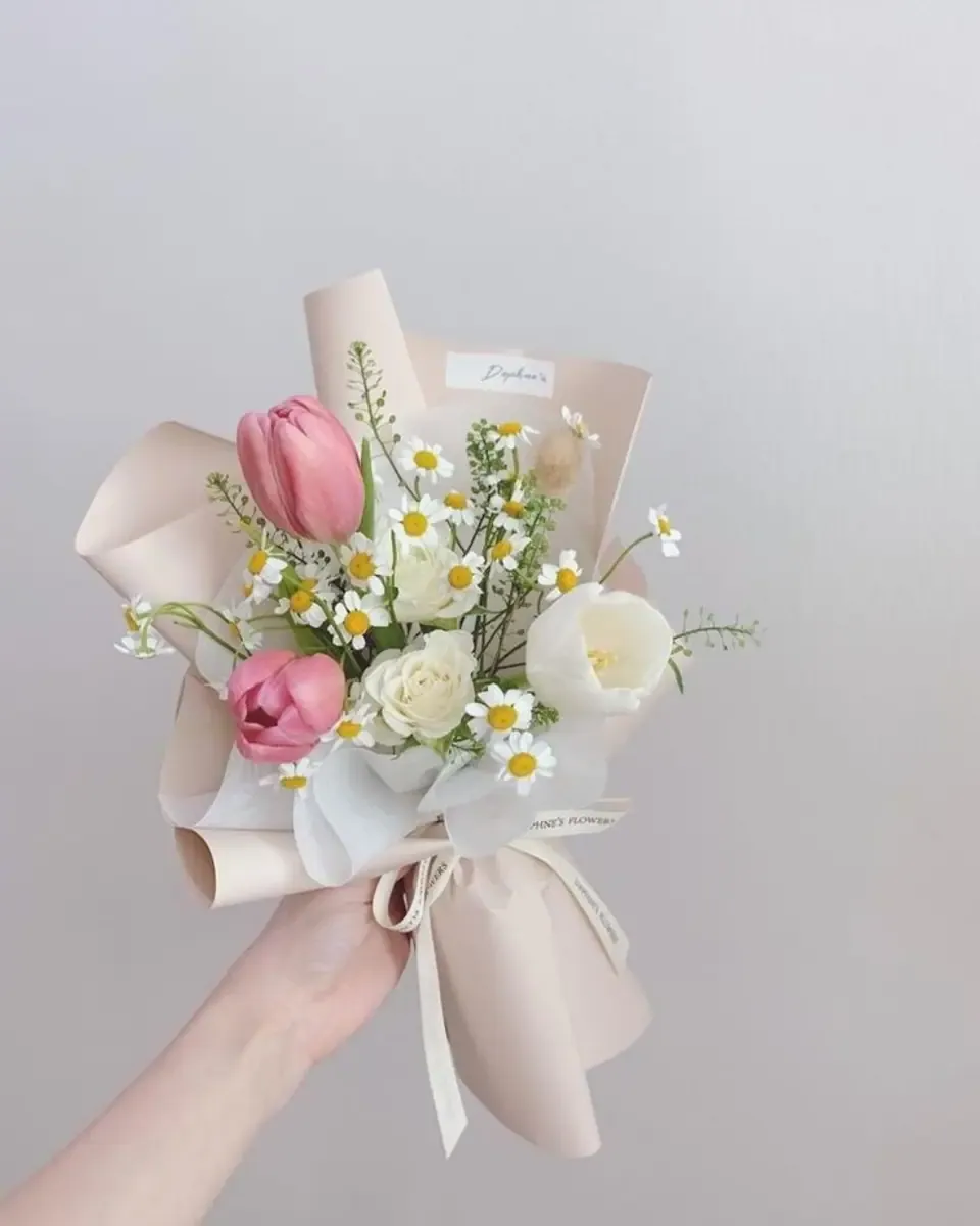Bó tulip hồng và cúc nhỏ gói giấy hồng nhạt, một tay cầm trước nền sáng