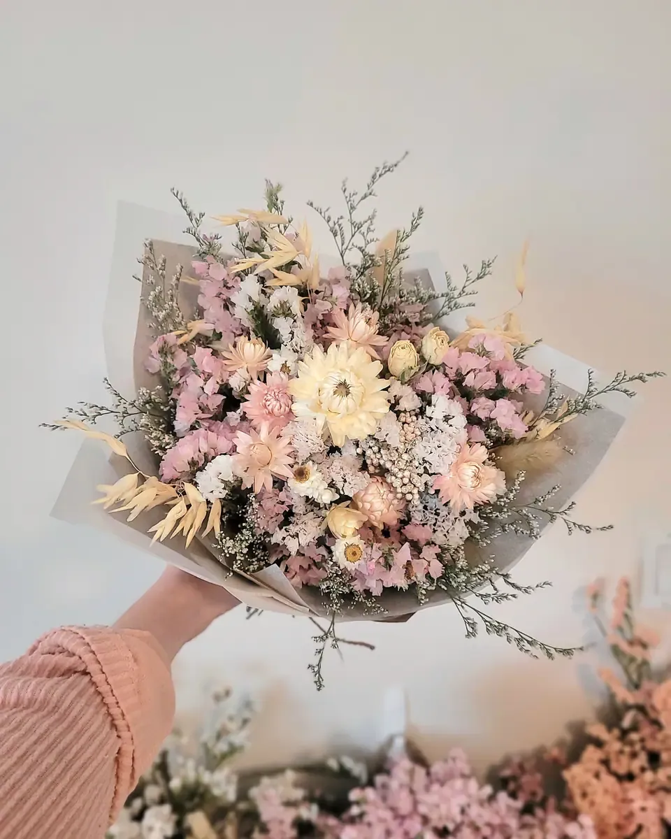 Bó hoa khô tông hồng kem được gói giấy mờ, một bàn tay nâng lên trước bức tường sáng