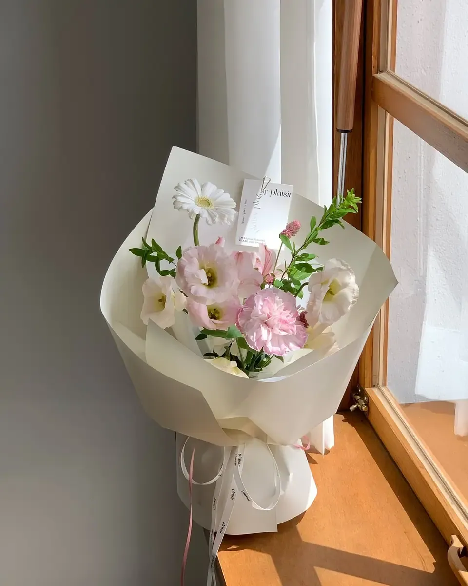 Bó hoa nhỏ gói giấy kem, hoa lisianthus hồng và cúc trắng buộc ruy băng, đặt cạnh cửa sổ gỗ