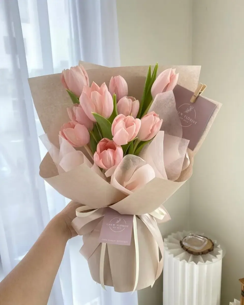 Bó tulip hồng gói giấy be và hồng nhạt, cầm tay cạnh cửa sổ sáng