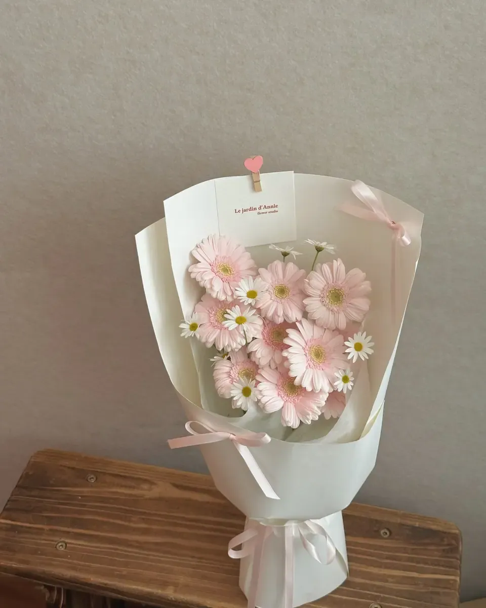 Bó hoa đồng tiền hồng nhạt cùng hoa cúc nhỏ gói giấy trắng, buộc nơ hồng và kẹp trái tim