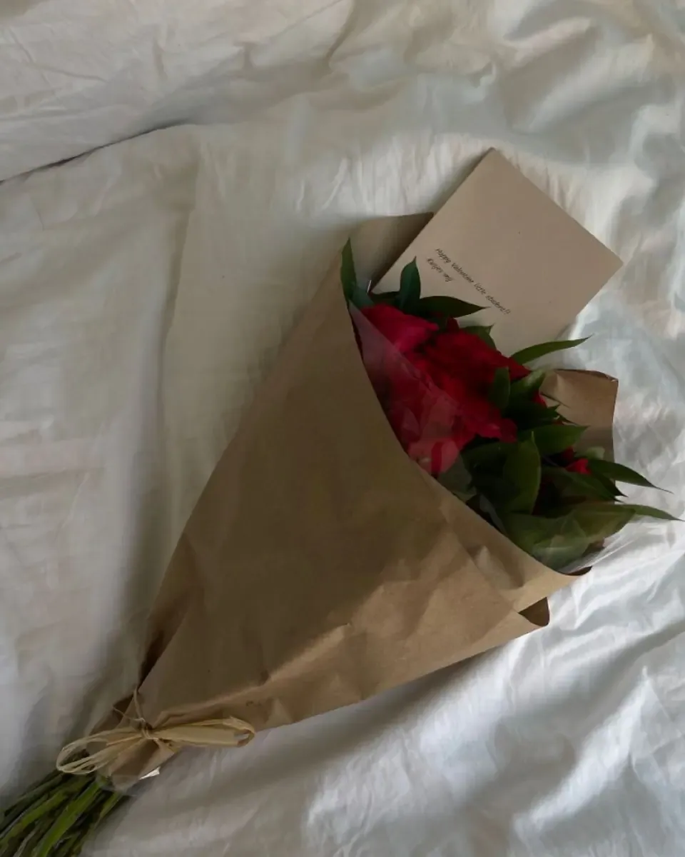 Bó hồng đỏ gói giấy kraft nằm trên ga trắng cùng tấm thiệp nhỏ