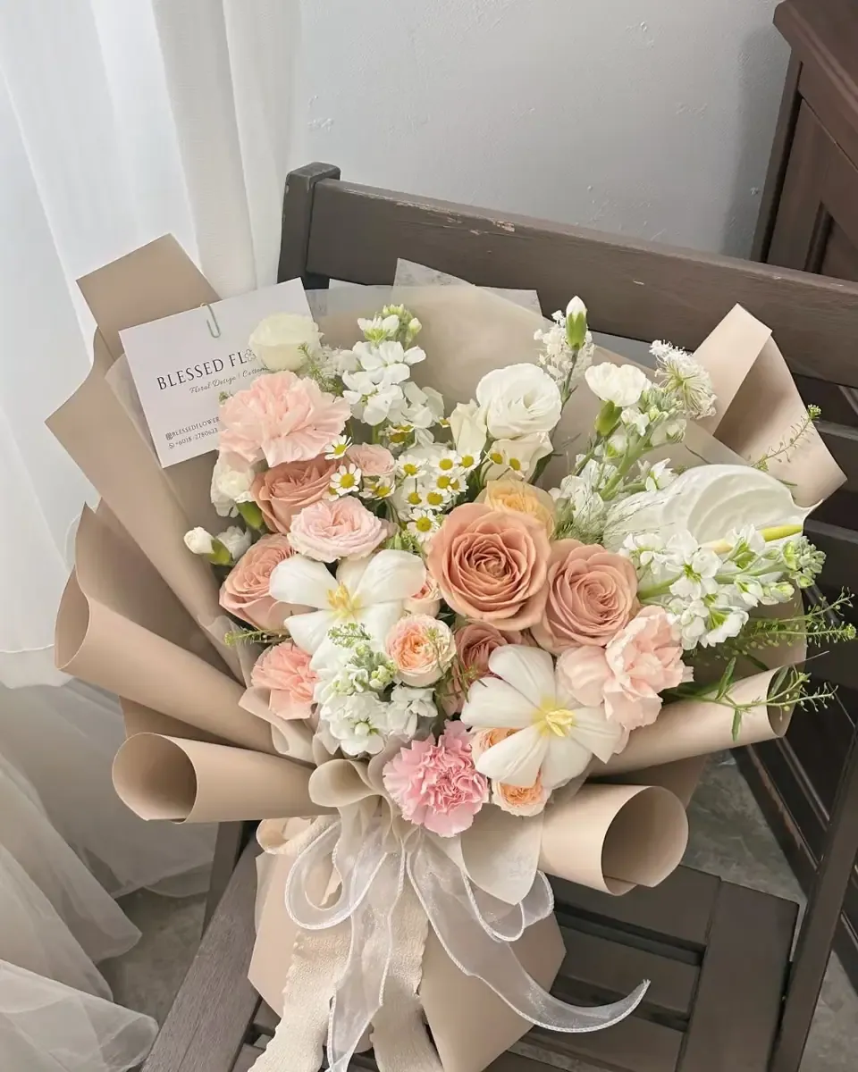 Bó hoa hồng, cẩm chướng và hoa trắng gói giấy kraft be, kèm thiệp nhỏ, đặt trên ghế gỗ