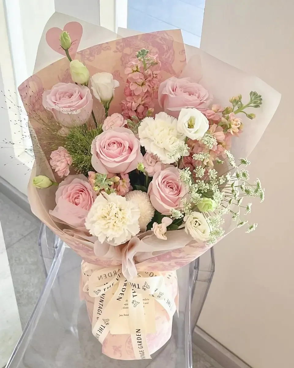 Bó hồng phấn, cẩm chướng trắng và hoa phụ gói giấy họa tiết hồng, đặt trên ghế trong