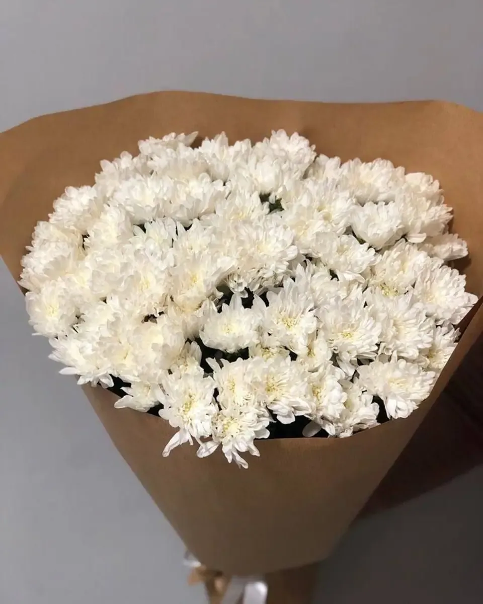 Bó cúc trắng nhiều bông gói giấy kraft nâu, dựng trước tường xám
