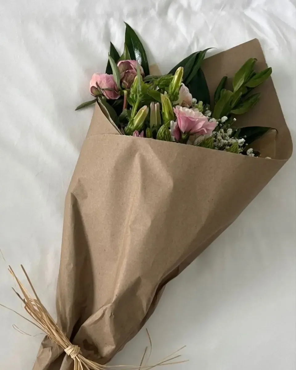 Bó hoa hồng, ly và lá xanh gói giấy kraft nằm trên ga giường trắng