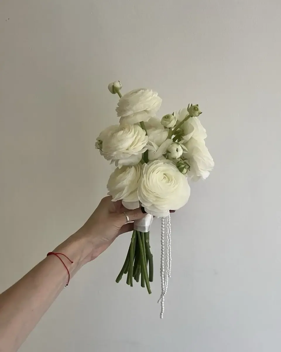 Bàn tay cầm bó hoa nhỏ gồm hoa mao lương trắng và lá xanh, cuống buộc ruy băng mỏng
