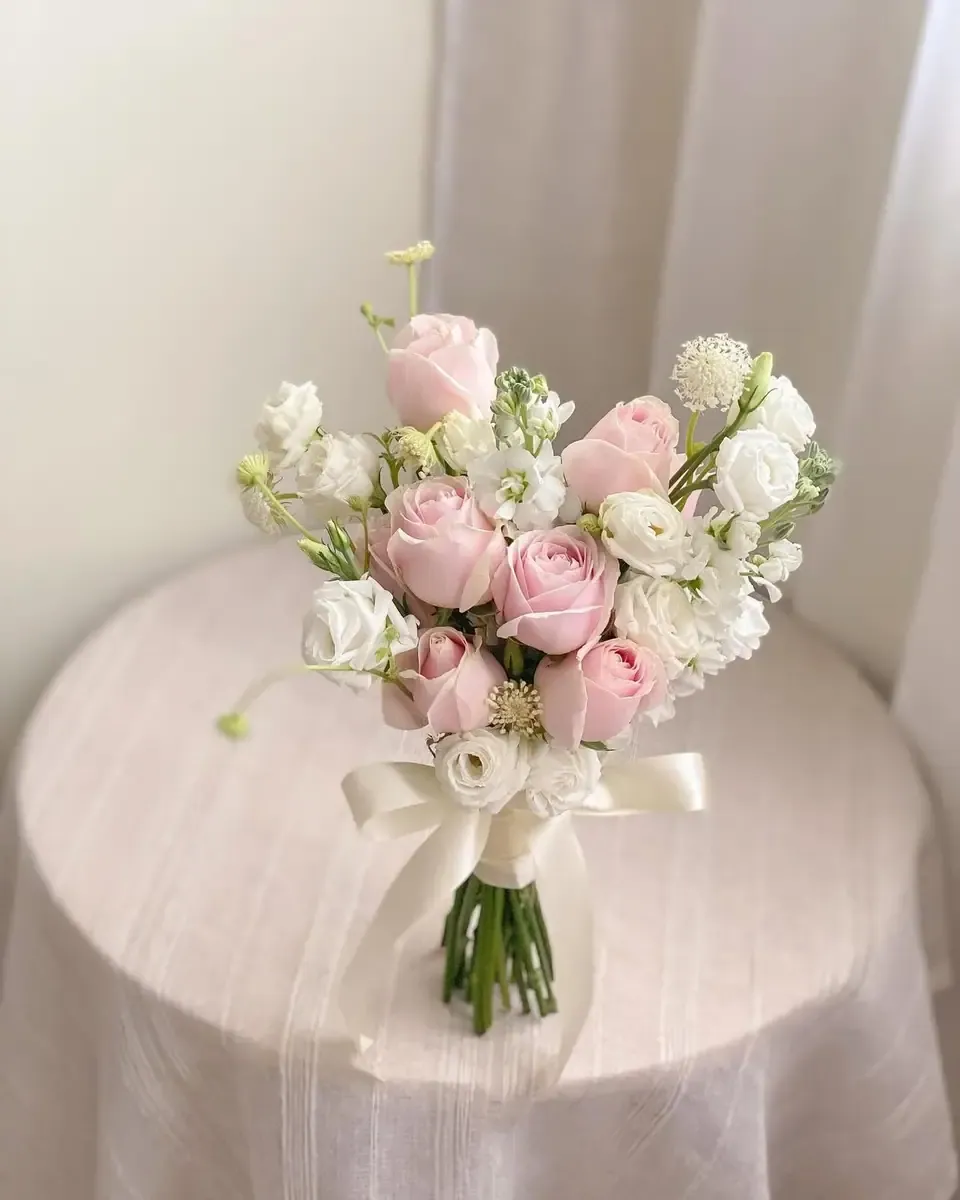 Bó hoa hồng hồng phấn, hoa trắng nhỏ buộc ruy băng kem đặt trên chiếc bàn tròn phủ khăn hồng