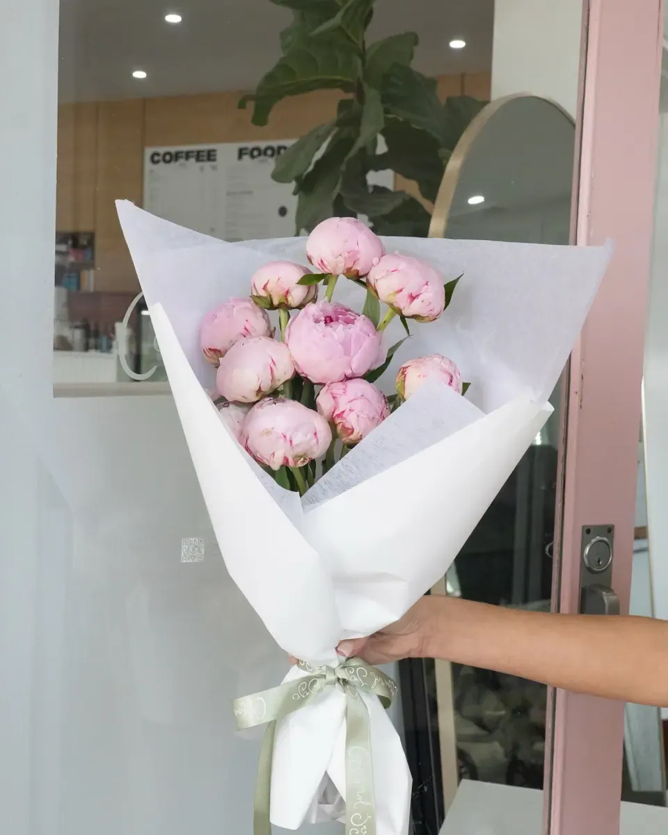 Bó mẫu đơn hồng nhạt gói giấy trắng, buộc ruy băng xanh lá, được cầm trước cửa kính