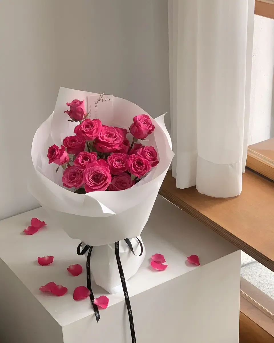 Bó hoa hồng hồng gói giấy trắng, rải cánh hoa trên bục trắng cạnh rèm cửa