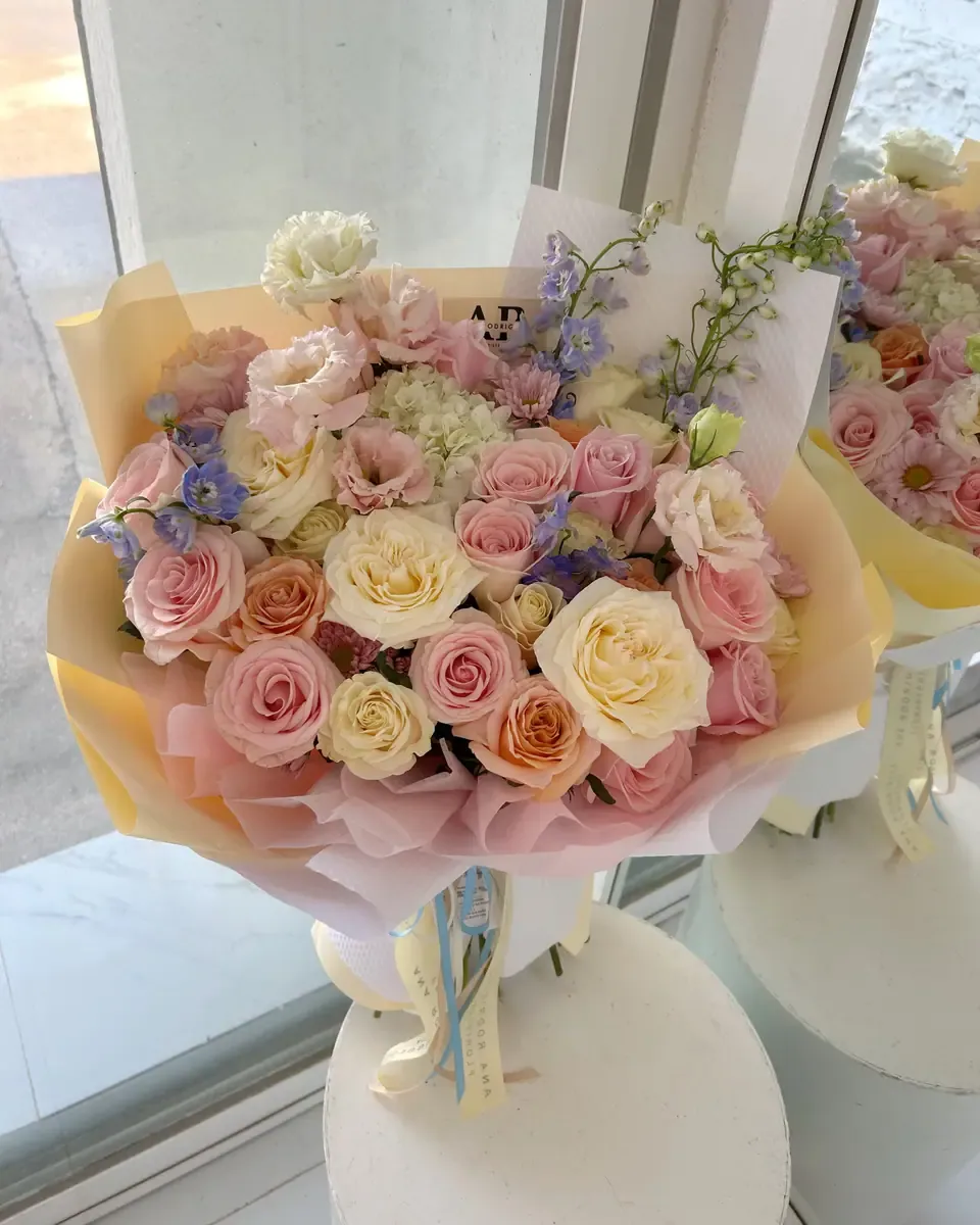 Bó hoa hồng đào, hoa kem và cúc gói giấy cam vàng, đặt cạnh cửa sổ có nắng