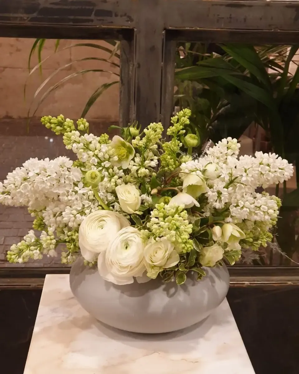 Chậu sứ xám thấp cắm hoa tử đinh hương trắng, hoa mao lương và lá xanh trên bệ đá cẩm thạch