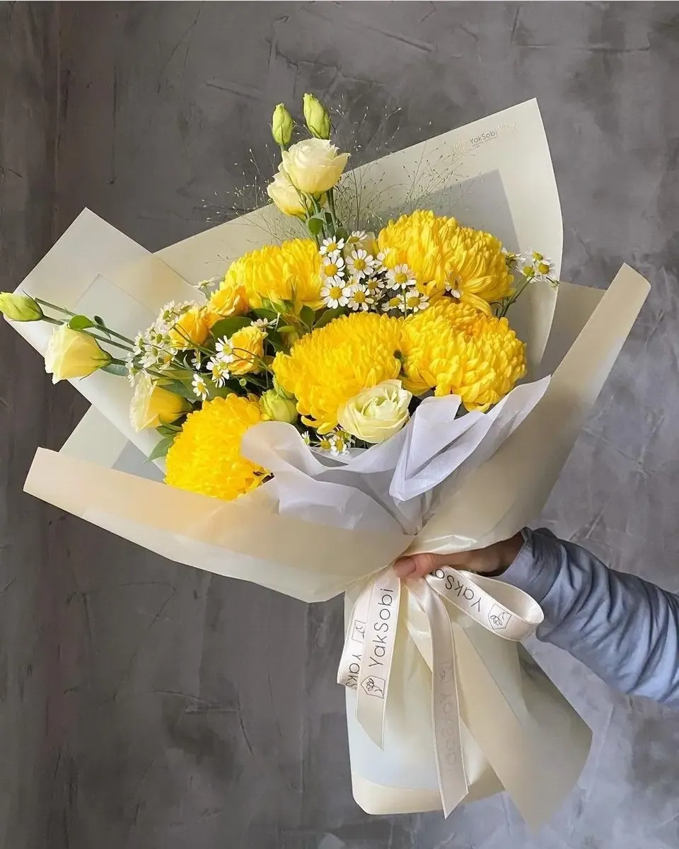 Bó hoa cúc vàng lớn kết hợp hoa kem gói giấy trắng, cầm trước tường xám