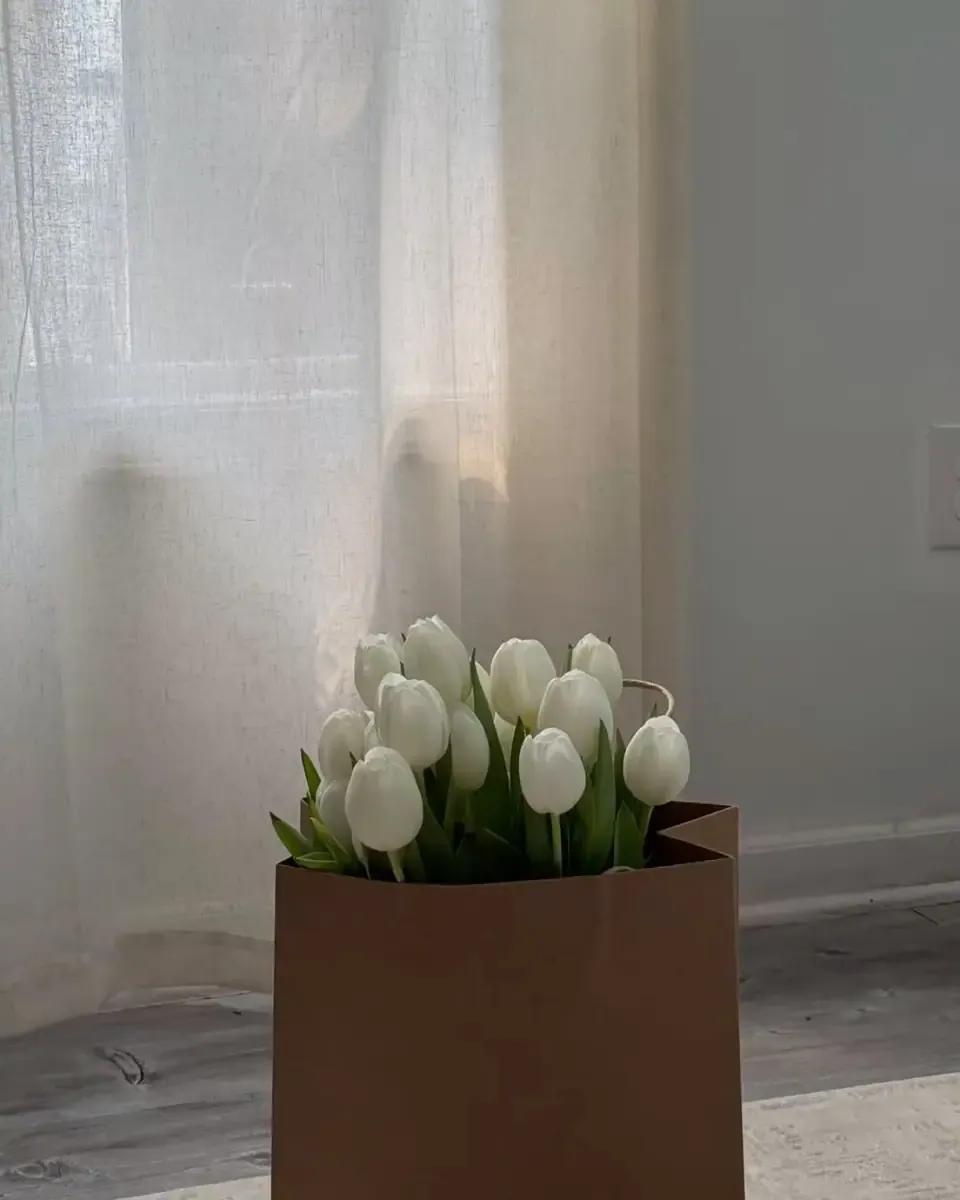 Một bó tulip trắng đặt trong túi giấy nâu cạnh tấm rèm mỏng, ánh sáng cửa sổ dịu