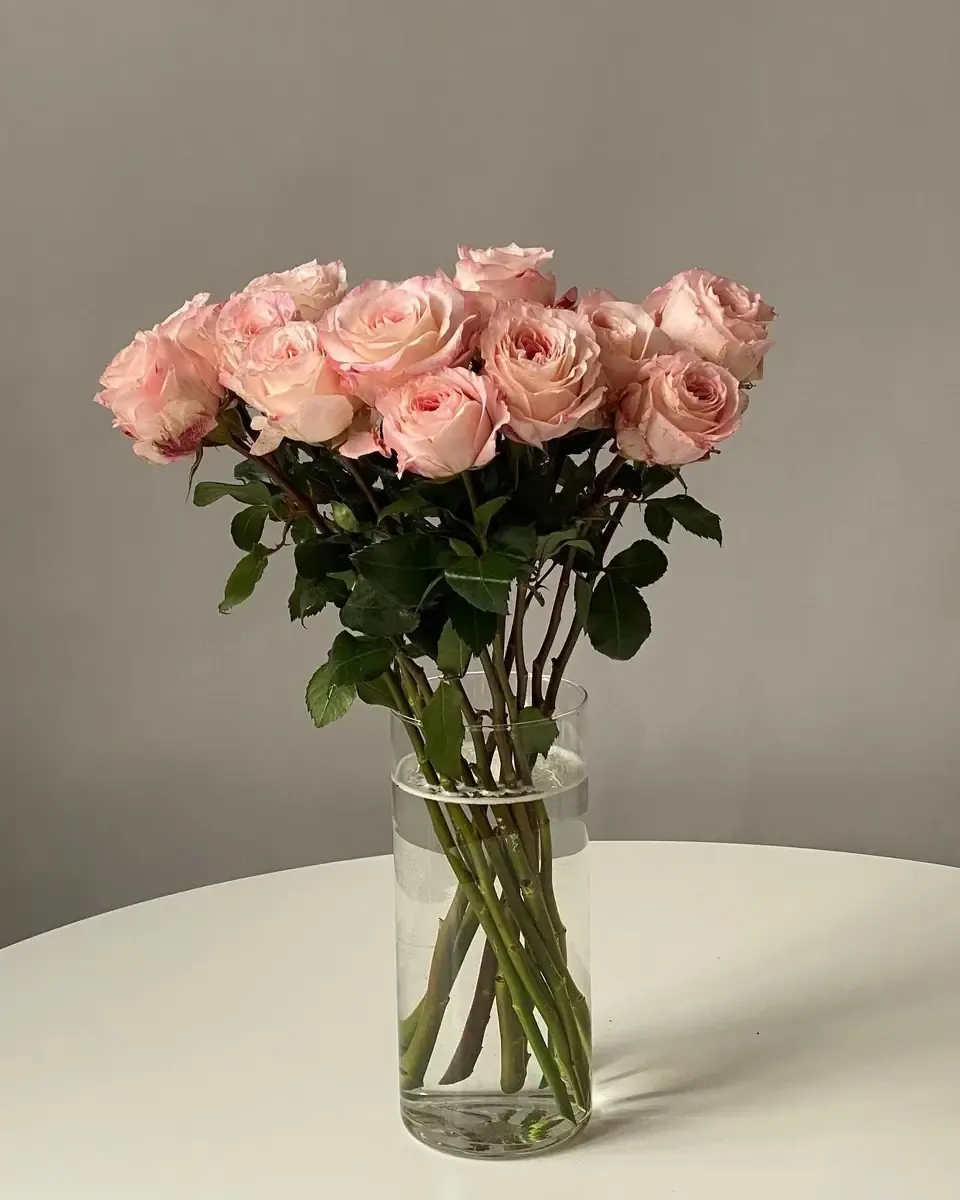 Bình thủy tinh có hoa hồng hồng đặt trên bàn tròn trắng trước nền tường xám