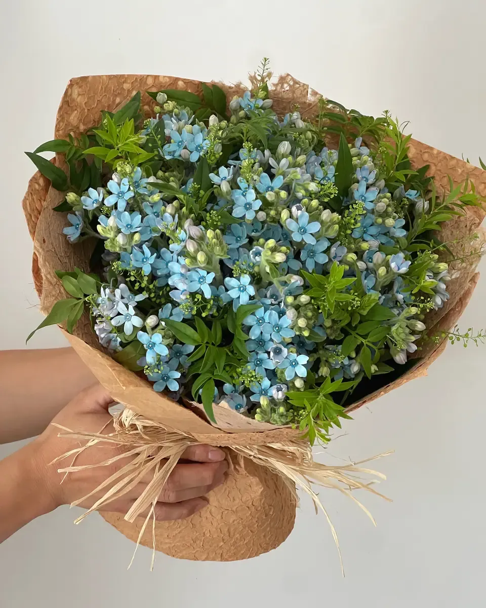 Bó hoa nhỏ màu xanh dương nhạt gói giấy kraft buộc dây rơm, một bàn tay cầm bó hoa