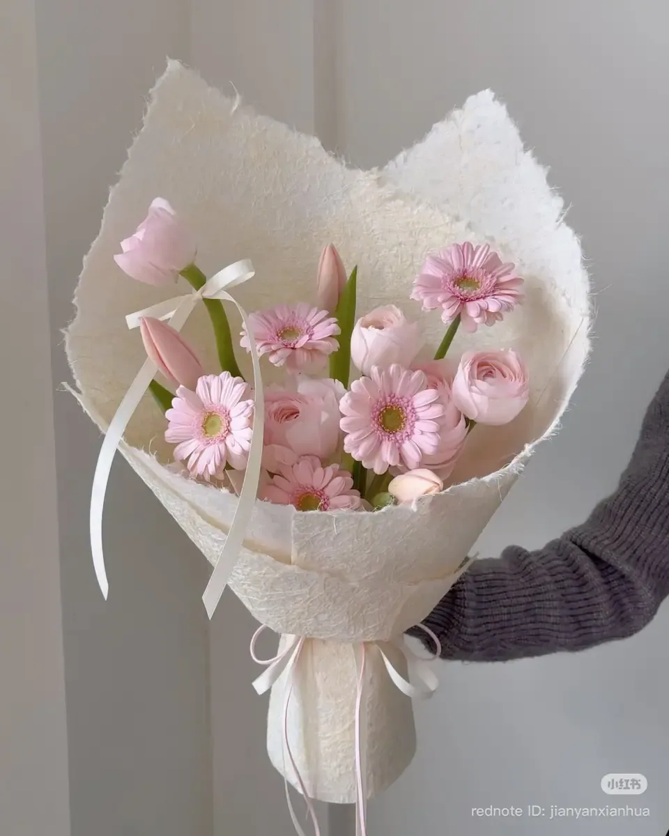 Bó hoa đồng tiền, hoa mao lương và tulip hồng gói giấy trắng thô, một tay cầm