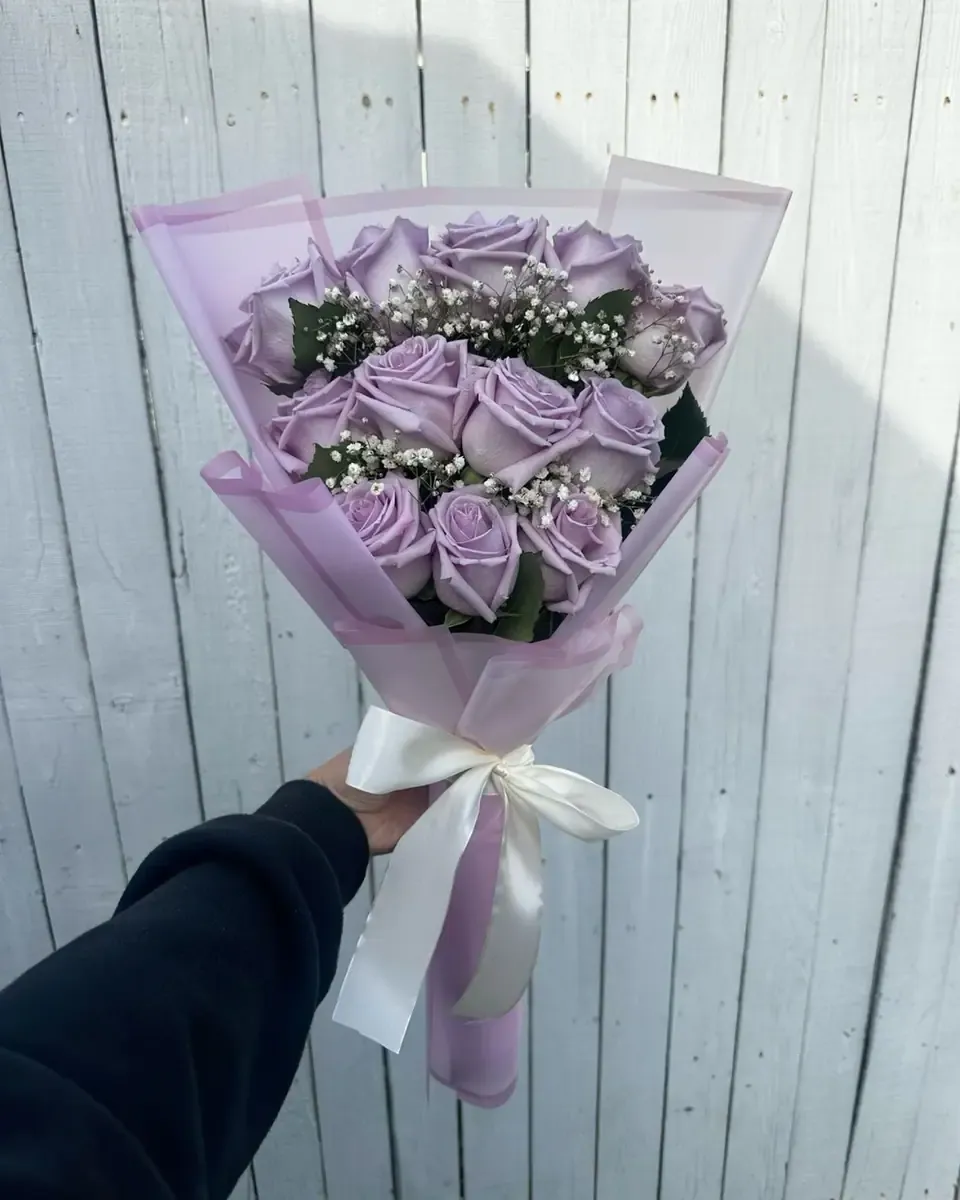 Bó hồng tím gói giấy tím nhạt, ruy băng trắng được cầm trước hàng rào trắng