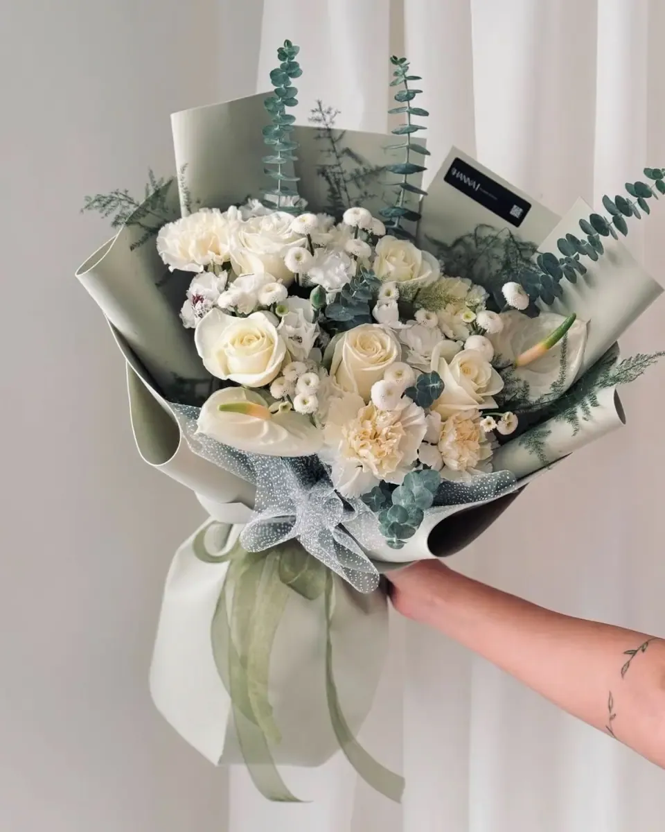 Bó hoa hồng trắng, cẩm chướng kem và lá bạch đàn gói giấy xanh sage, được tay cầm