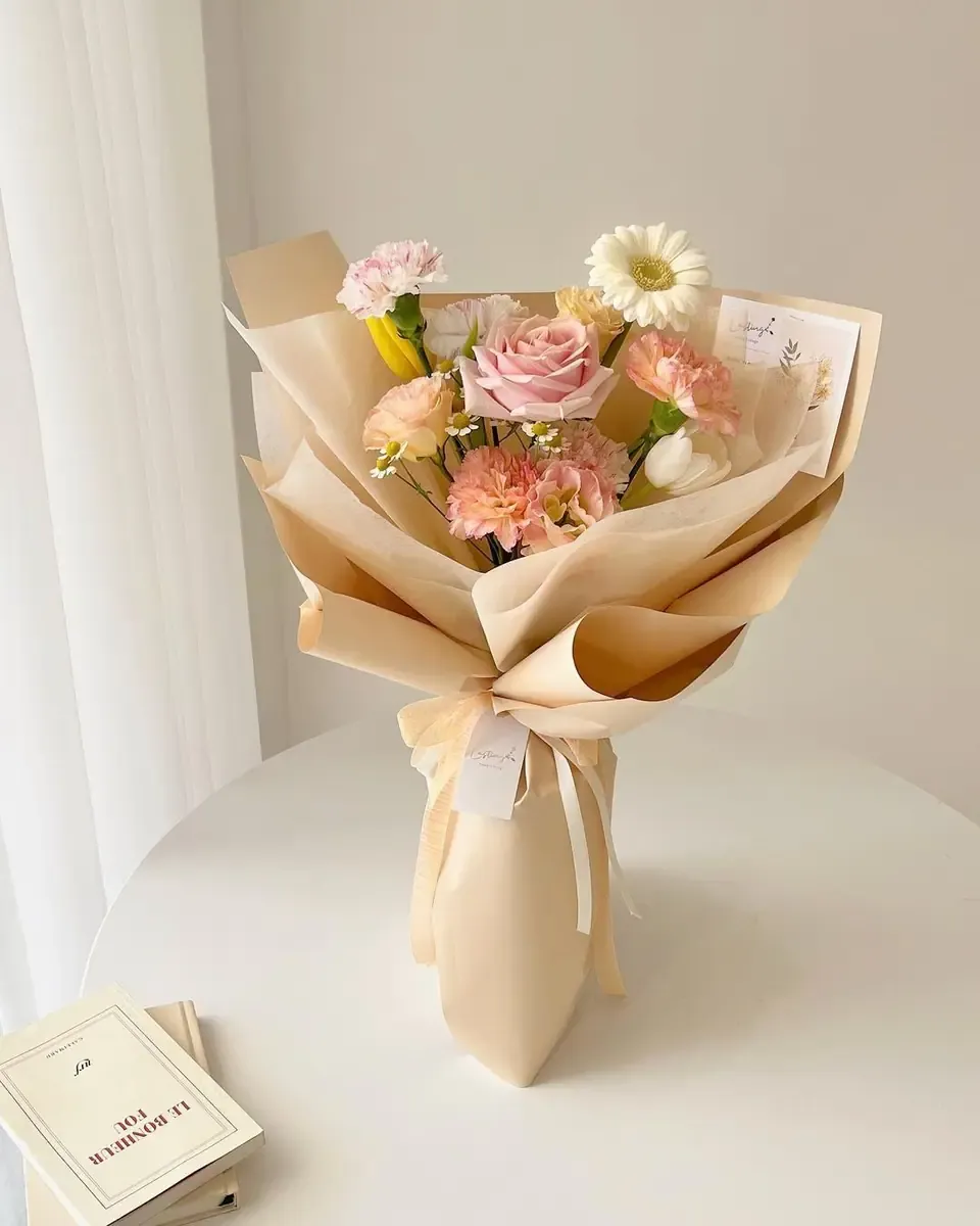 Bó hoa tông hồng đào gói giấy kem, buộc ruy băng màu be, đặt trên bàn trắng cạnh cuốn sách