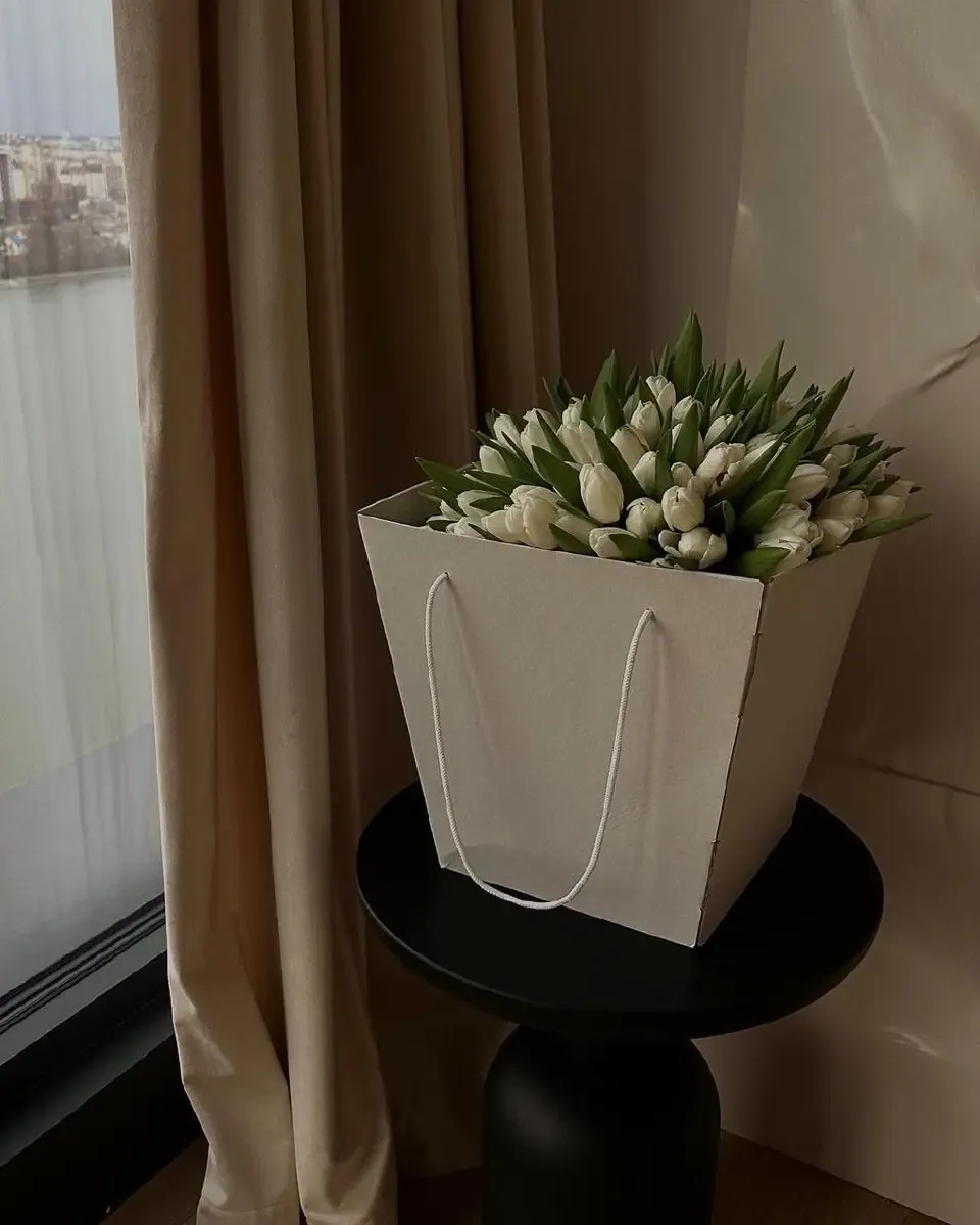 Túi giấy trắng có quai cầm đựng bó tulip trắng, đặt trên bàn tròn đen cạnh rèm be