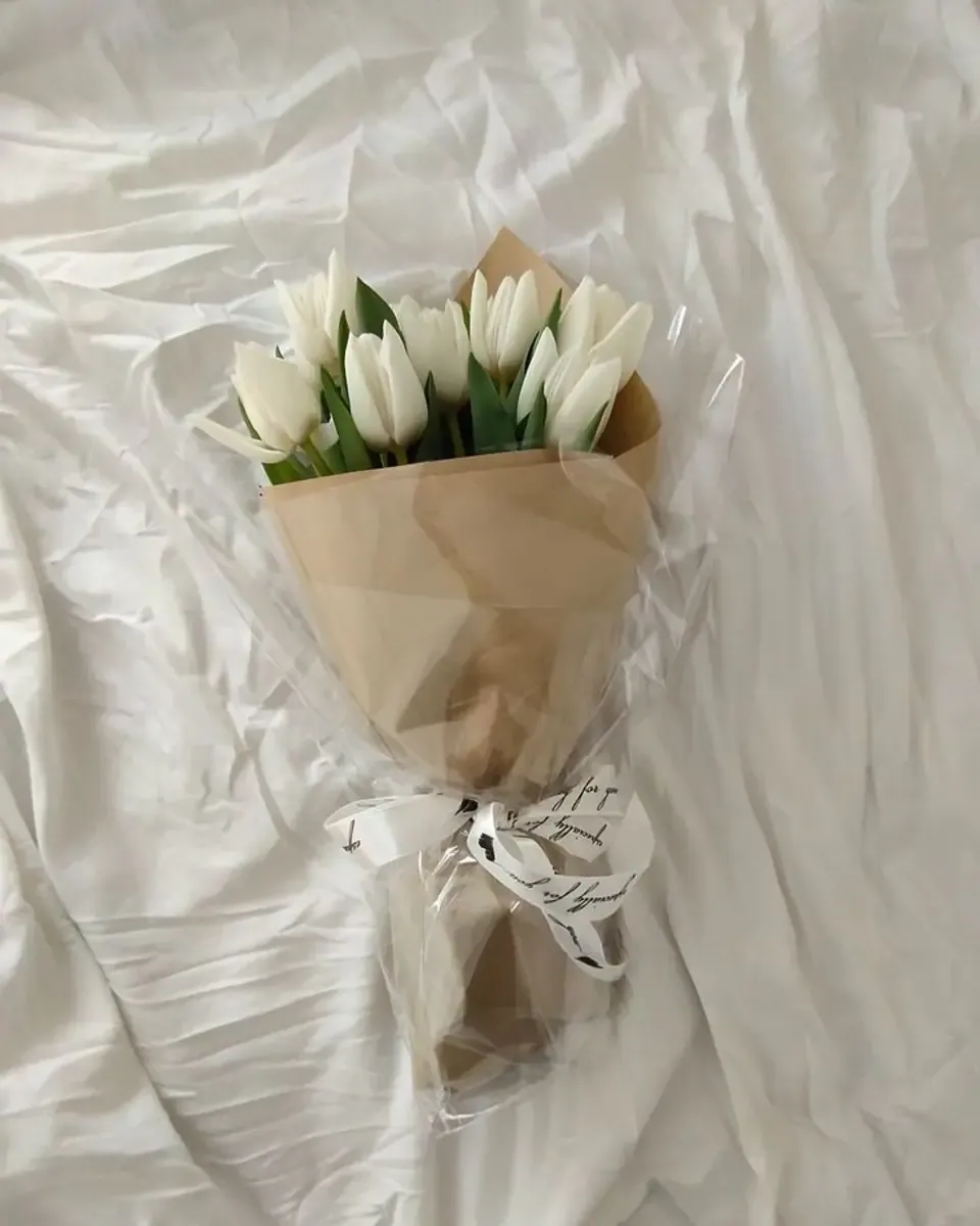 Bó tulip trắng gói giấy kraft và bọc trong suốt, buộc nơ trắng, đặt trên ga giường trắng
