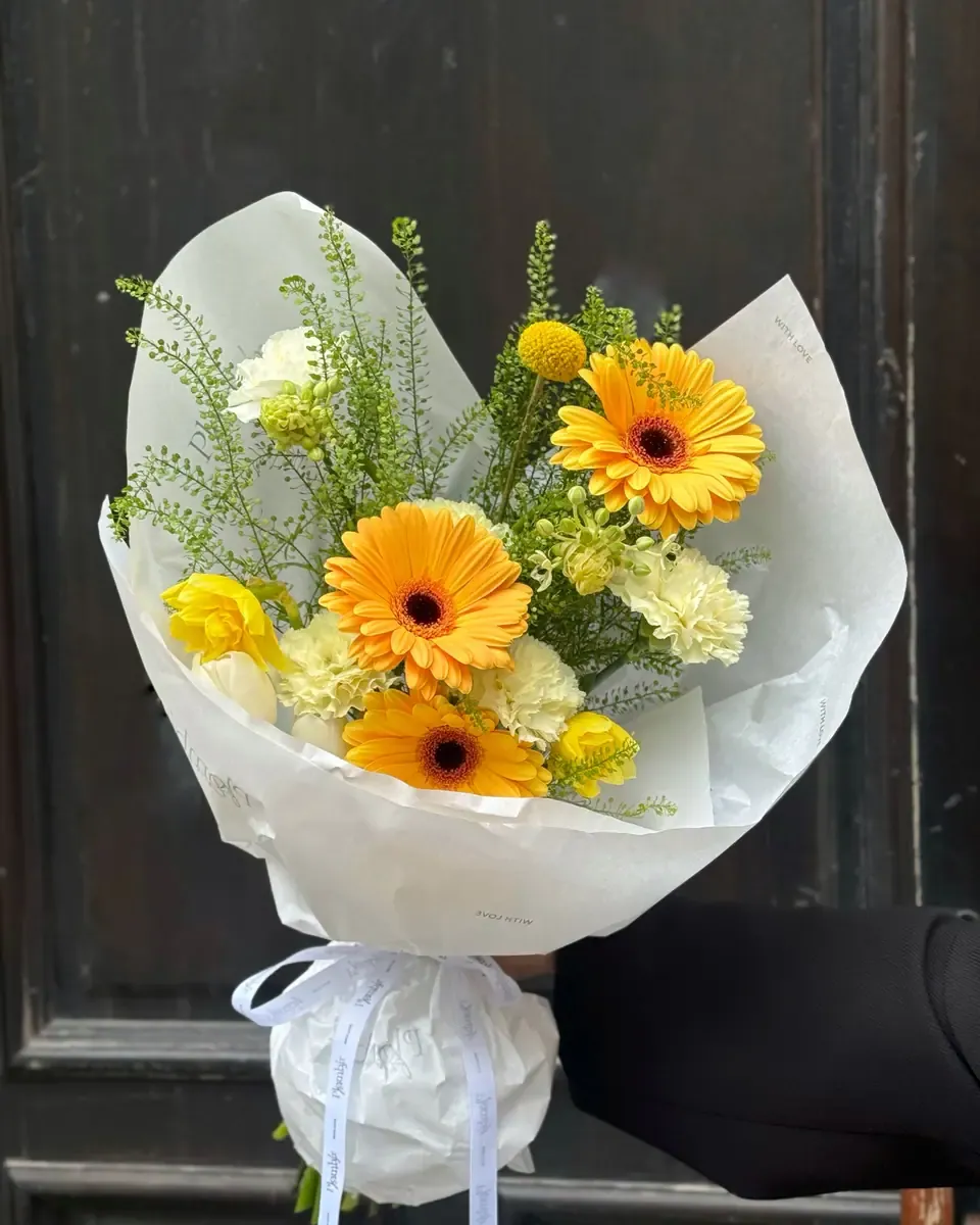 Bó gerbera vàng cam cùng lá xanh gói giấy trắng đặt trong quán