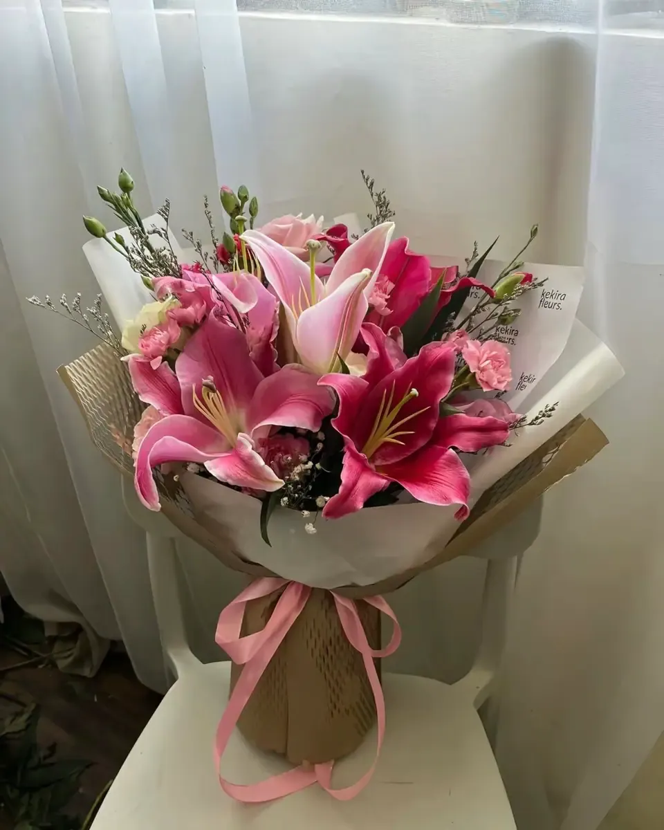 Bó ly hồng đậm gói giấy trắng và kraft, buộc ruy băng hồng cạnh rèm trắng