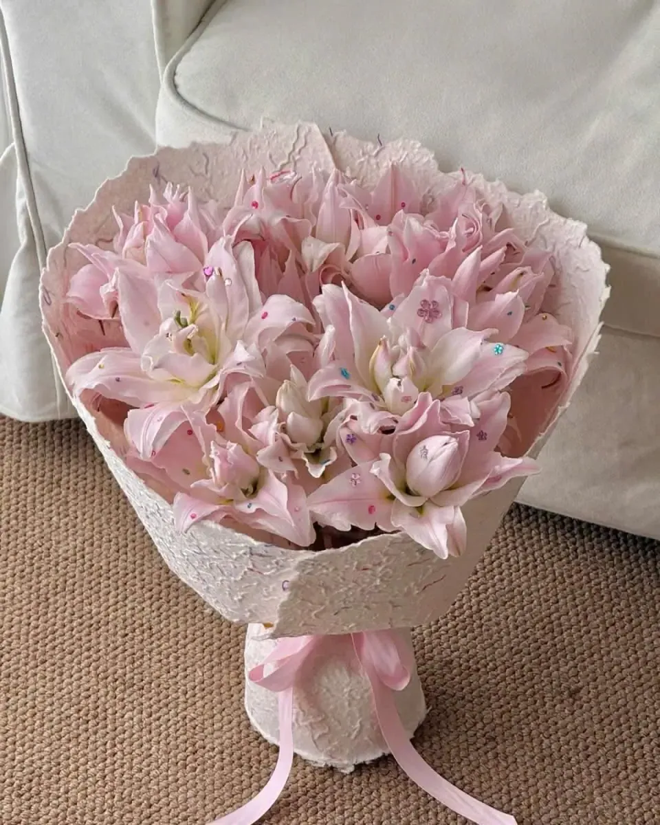 Bó hoa ly hồng gói giấy ren trắng, buộc nơ hồng, đặt trên thảm sợi màu be