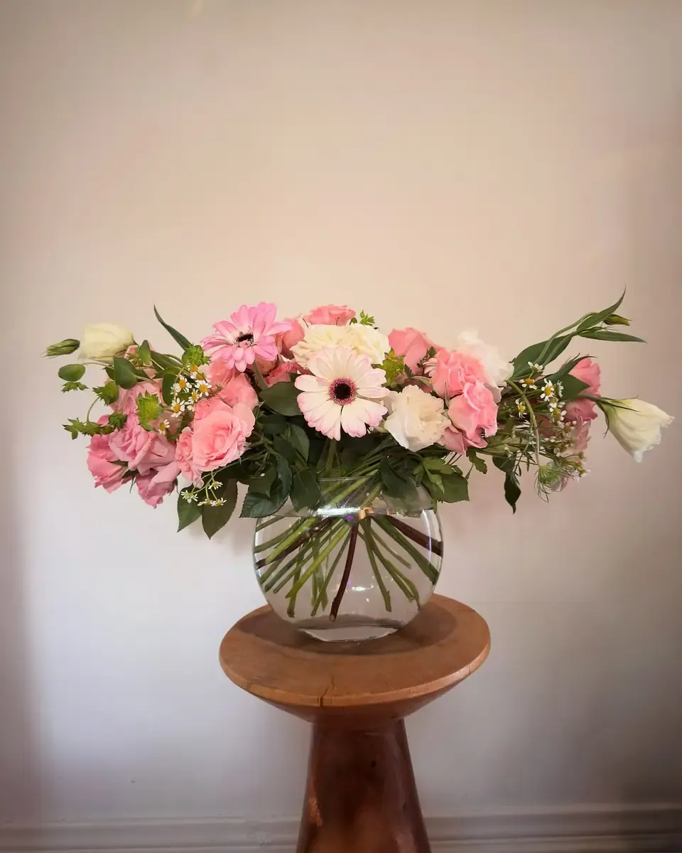 Hoa gerbera hồng, cẩm chướng và lá xanh trong bình thủy tinh tròn đặt trên ghế gỗ nâu