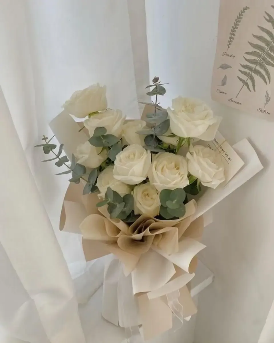 Bó hoa hồng trắng cài lá bạch đàn gói giấy kem và kraft đặt gần cửa sổ