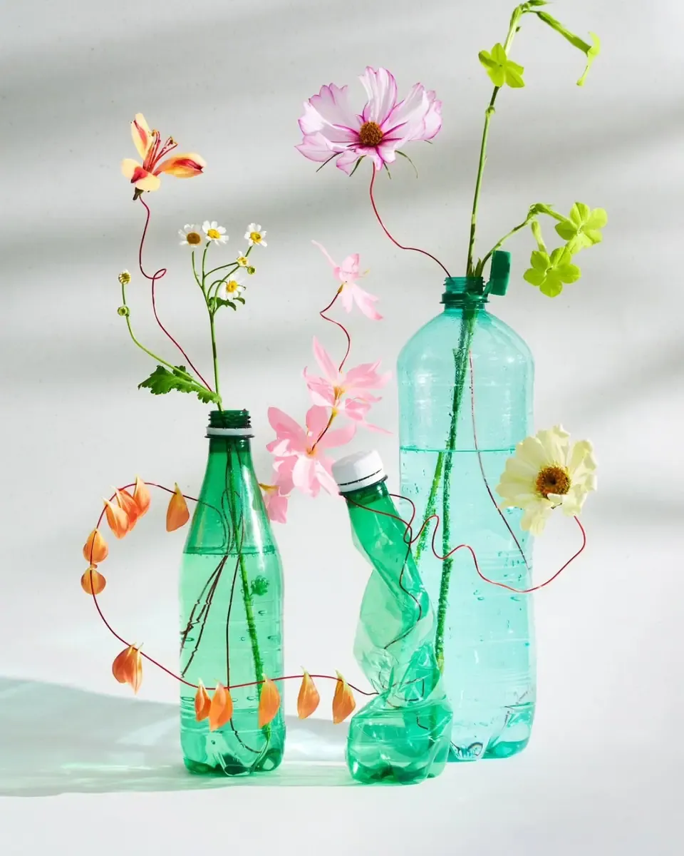 Hai chai nhựa xanh ngọc cắm hoa cosmos hồng và hoa nhỏ trên nền sáng có nắng