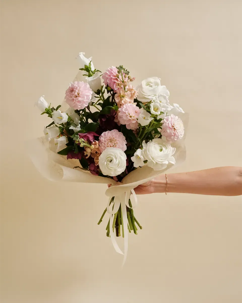 Bó hoa nhỏ hồng phấn, trắng và tím nhạt gói giấy kem, tay đưa ra