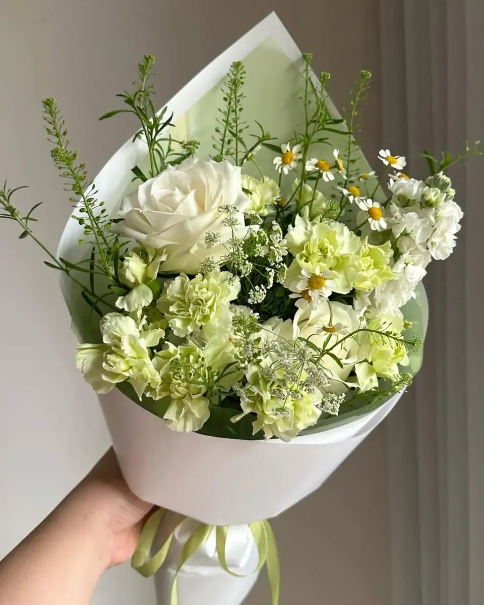 Bó hoa gói giấy trắng gồm hồng trắng, cẩm chướng xanh nhạt và cành lá mảnh, cầm trước rèm cửa
