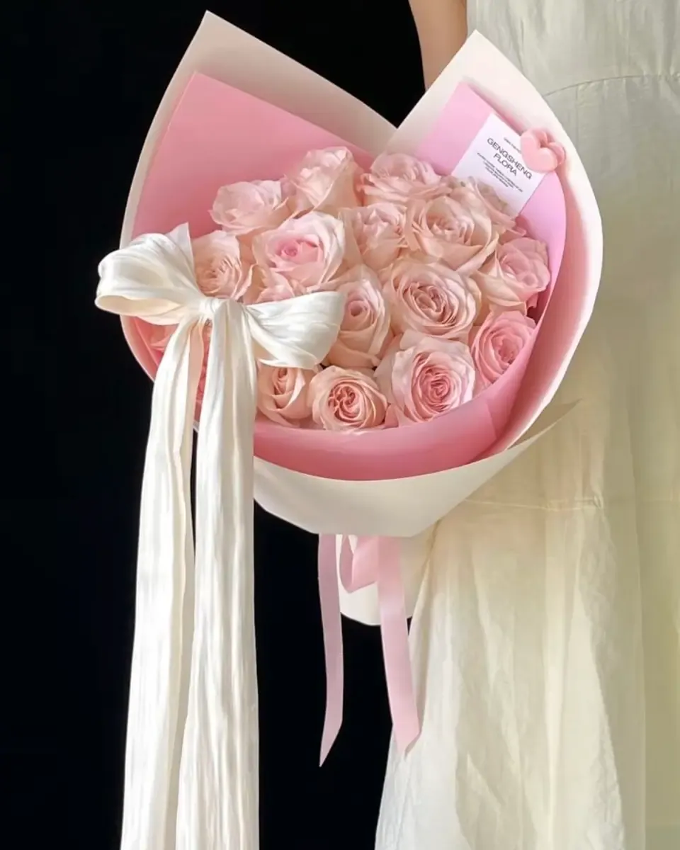 Bó hoa hồng phấn gói giấy hồng và kem, thắt ruy băng lụa trắng dài, một người mặc váy kem cầm trên tay