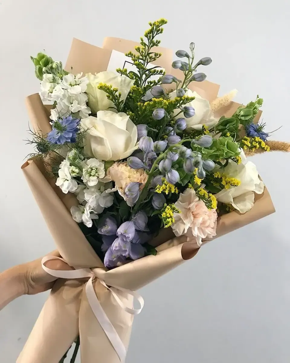 Bó hoa hồng trắng, hoa xanh tím và cỏ vàng gói giấy nâu kem, buộc ruy băng hồng nhạt