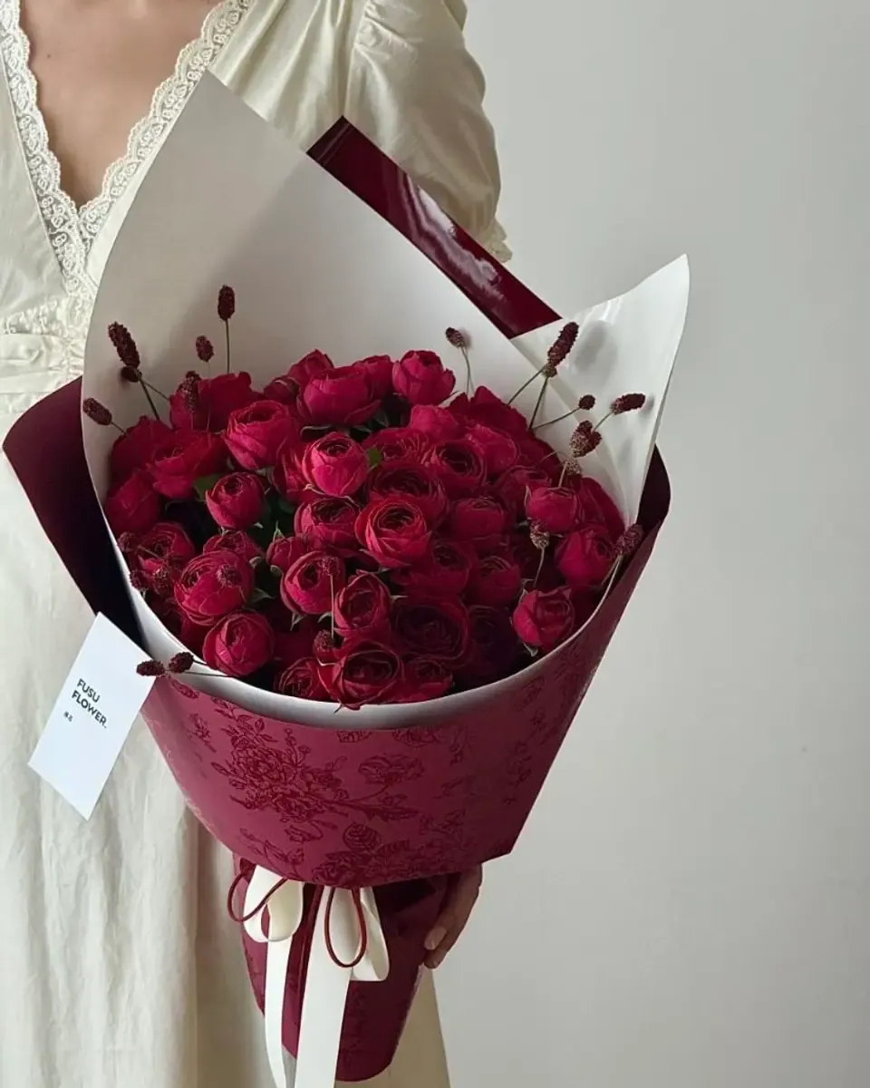 Bó hoa hồng đỏ sẫm gói giấy đỏ rượu, buộc ruy băng kem, cầm trên tay
