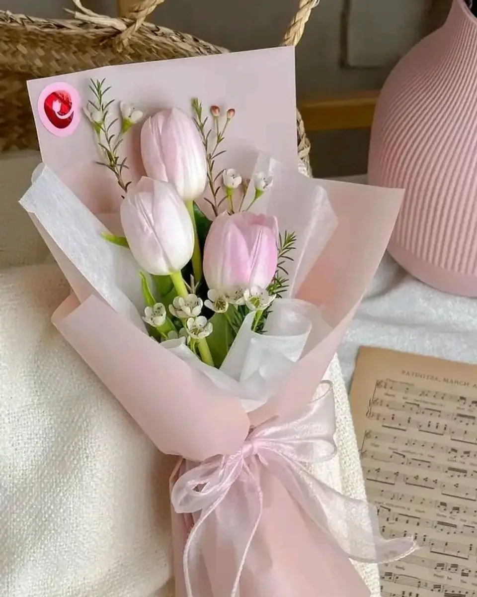Bó tulip hồng nhạt cùng hoa trắng nhỏ gói giấy hồng, buộc ruy băng voan, đặt cạnh tờ nhạc