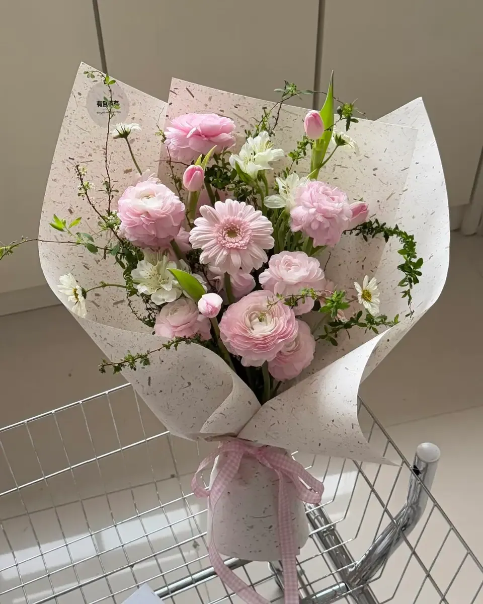 Bó hoa mao lương hồng, gerbera hồng và lá xanh gói giấy trắng lốm đốm, đặt cạnh giỏ lưới kim loại