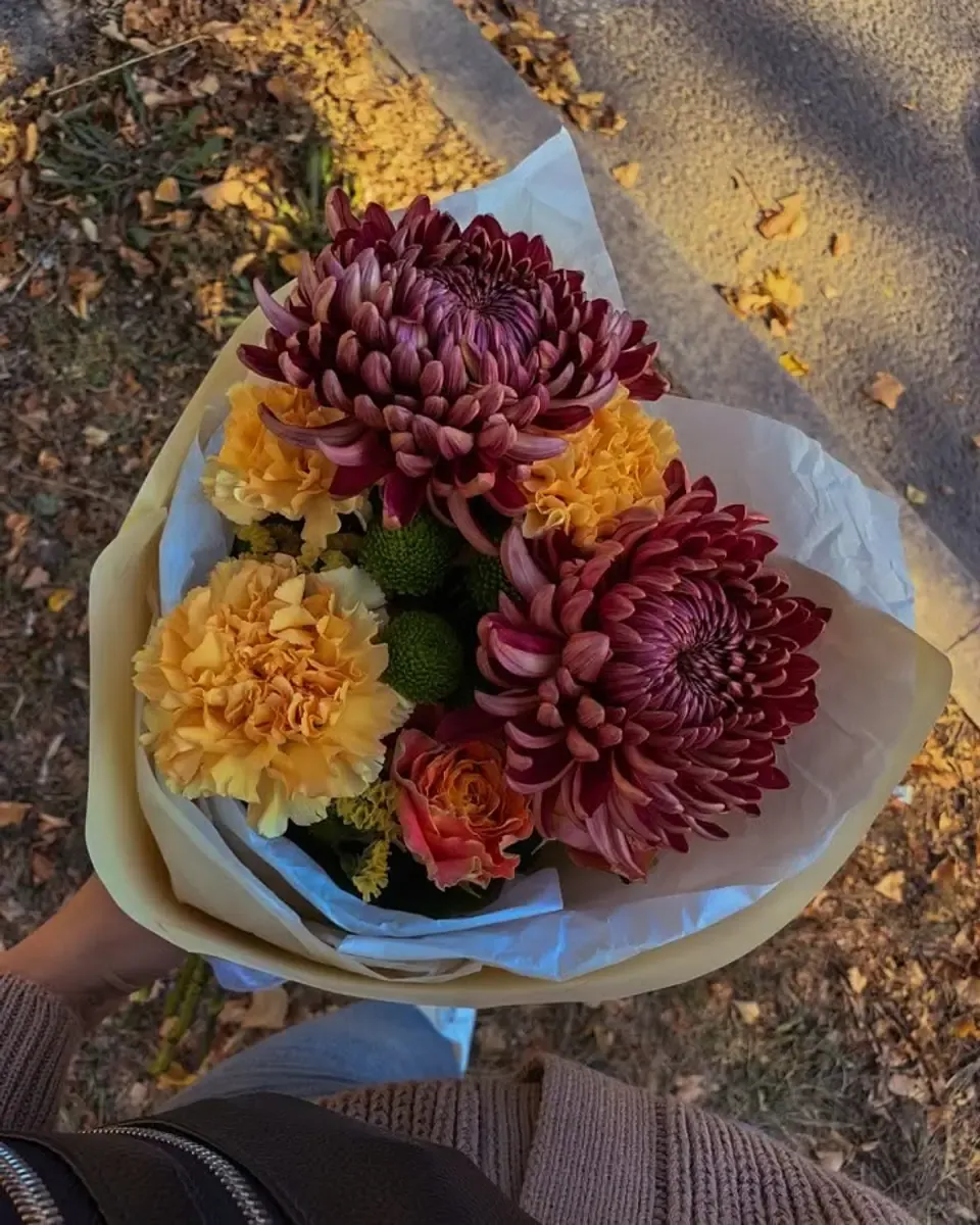 Bó cúc đại đóa đỏ đô và vàng gói giấy kem, cầm trên đường lá thu