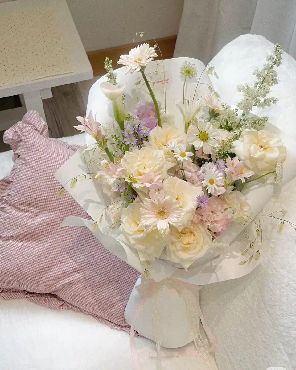 Bó hoa pastel kem, hồng nhạt và xanh dịu gói giấy trắng, đặt trên giường