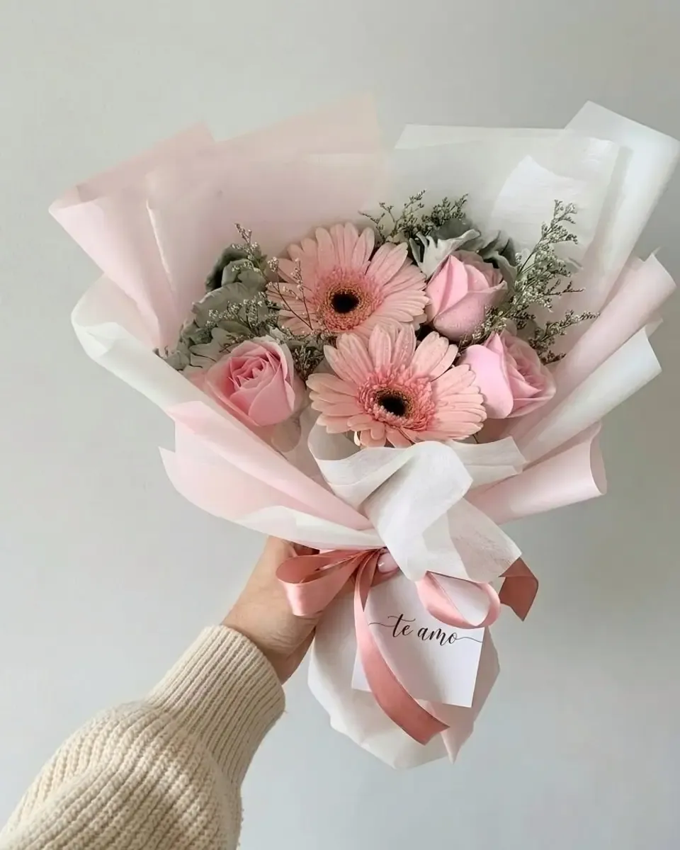 Bó hoa đồng tiền hồng, hồng bông nhỏ và lá xám gói giấy hồng và trắng, một tay cầm
