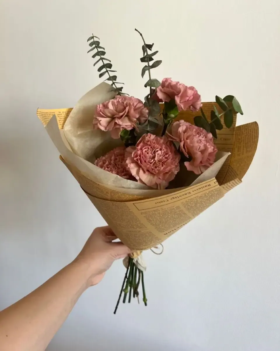 Bó cẩm chướng hồng đất gói giấy kraft báo, được cầm trên tay cạnh nền sáng