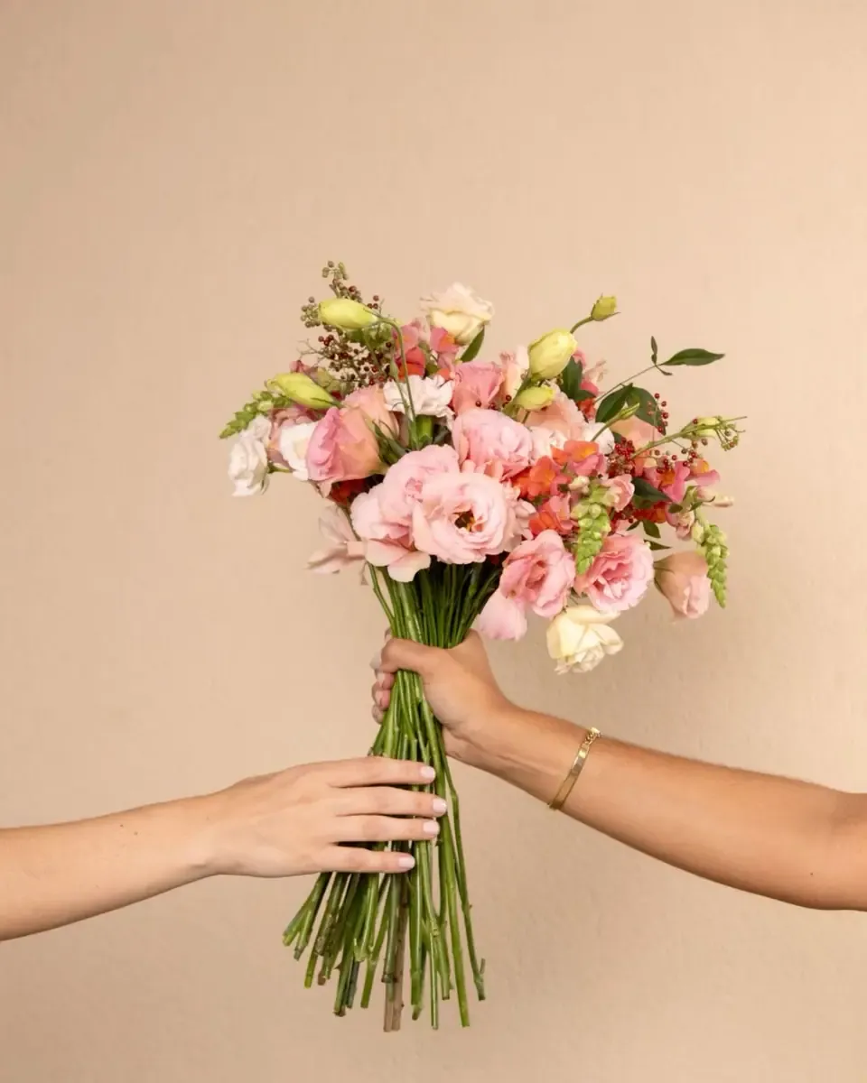 Bó hoa lisianthus hồng cam gói gọn, tay hai người trao bó hoa nền be